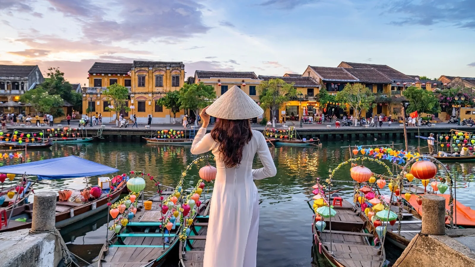 A woman in a white ao dai and conical hat admires the lantern-lit riverside boats of Hoi An at dusk. A woman in a white ao dai and conical hat admires the lantern-lit riverside boats of Hoi An at dusk.
