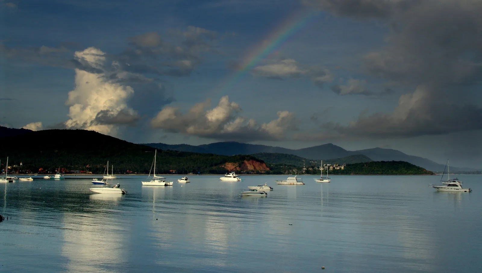A rainbow arcs over a calm bay dotted with boats and hills near Koh Samui, Thailand.